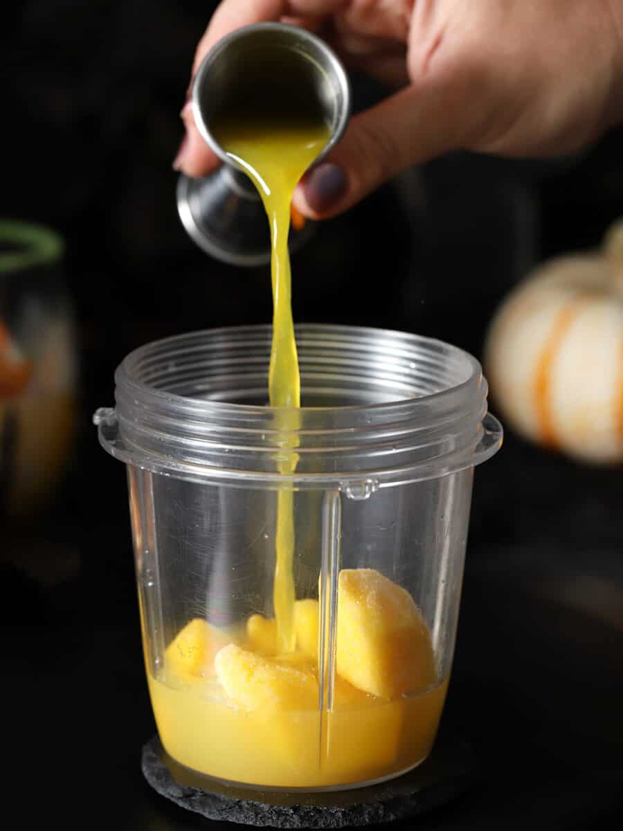 Orange juice being poured into a blender cup with mango and pineapple juice.