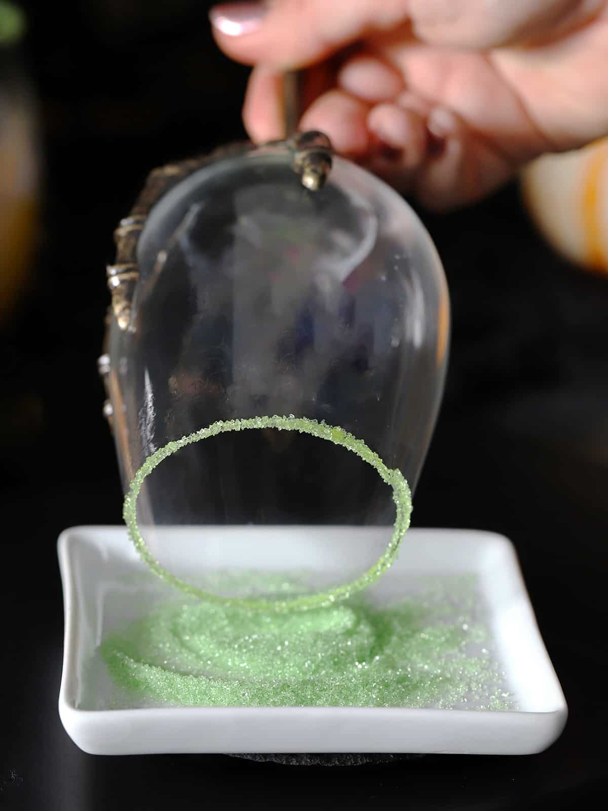 A hand dipping the rim of a glass into green sugar. 