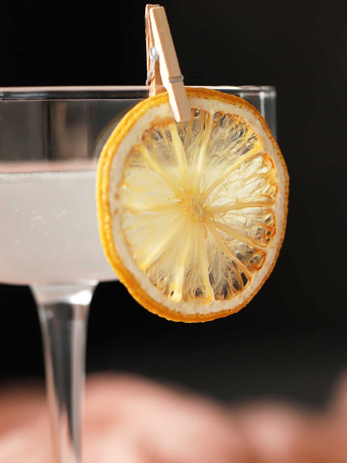 A piece of dried orange held on to the side of a cocktail glass with a tiny clothespin. 