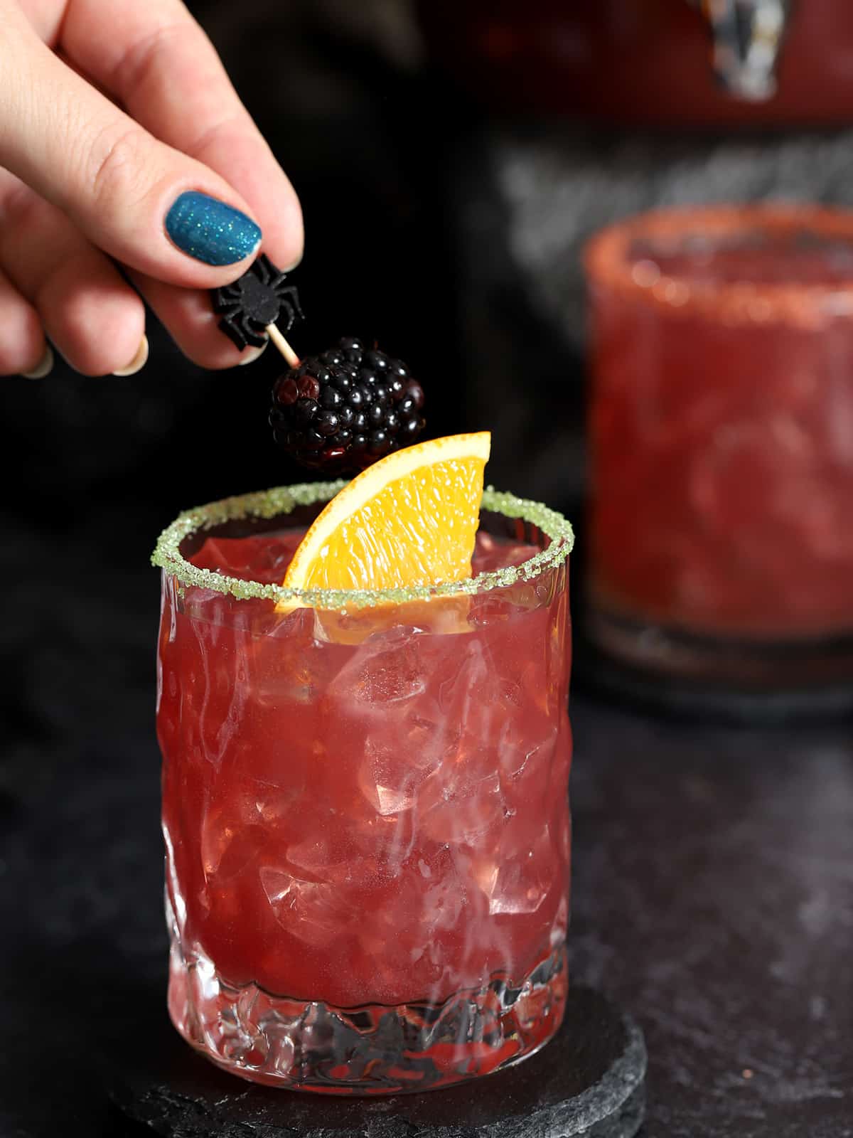 A hand placing a garnish pick with blackberry and orange on it into a glass of Halloween punch. 