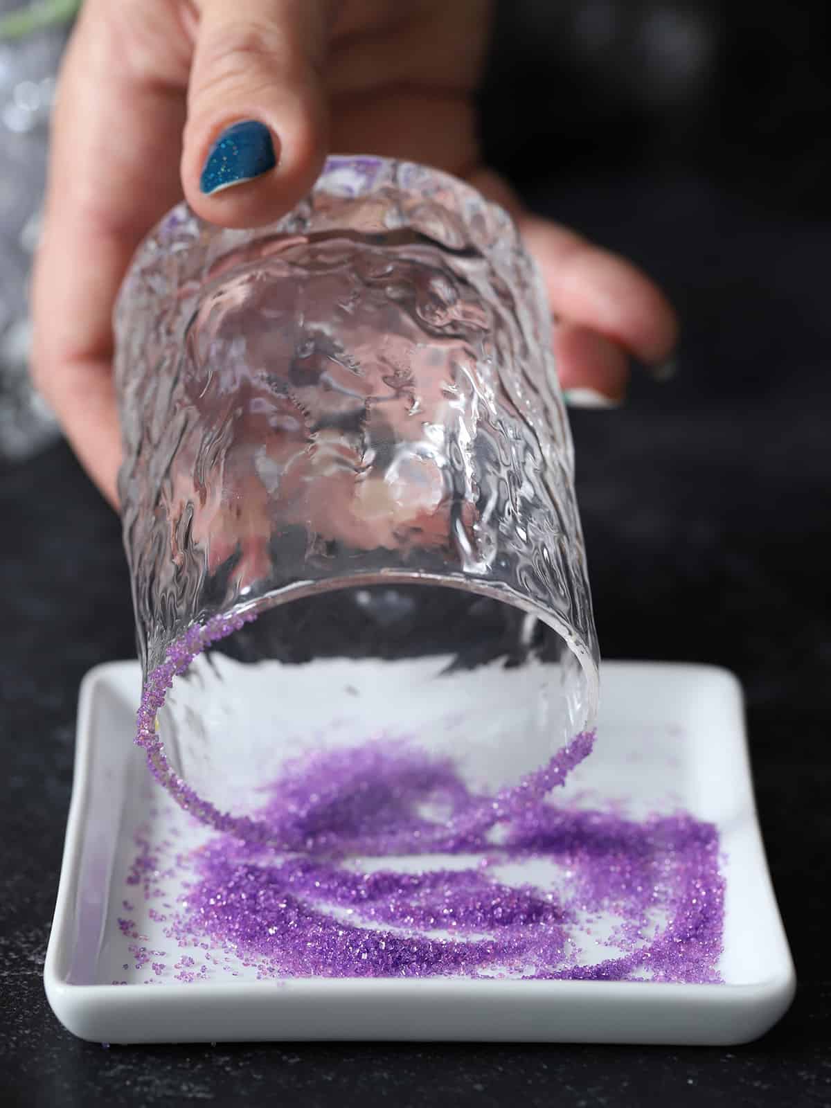 A hand dipping the rim of a glass into purple decorating sugar. 