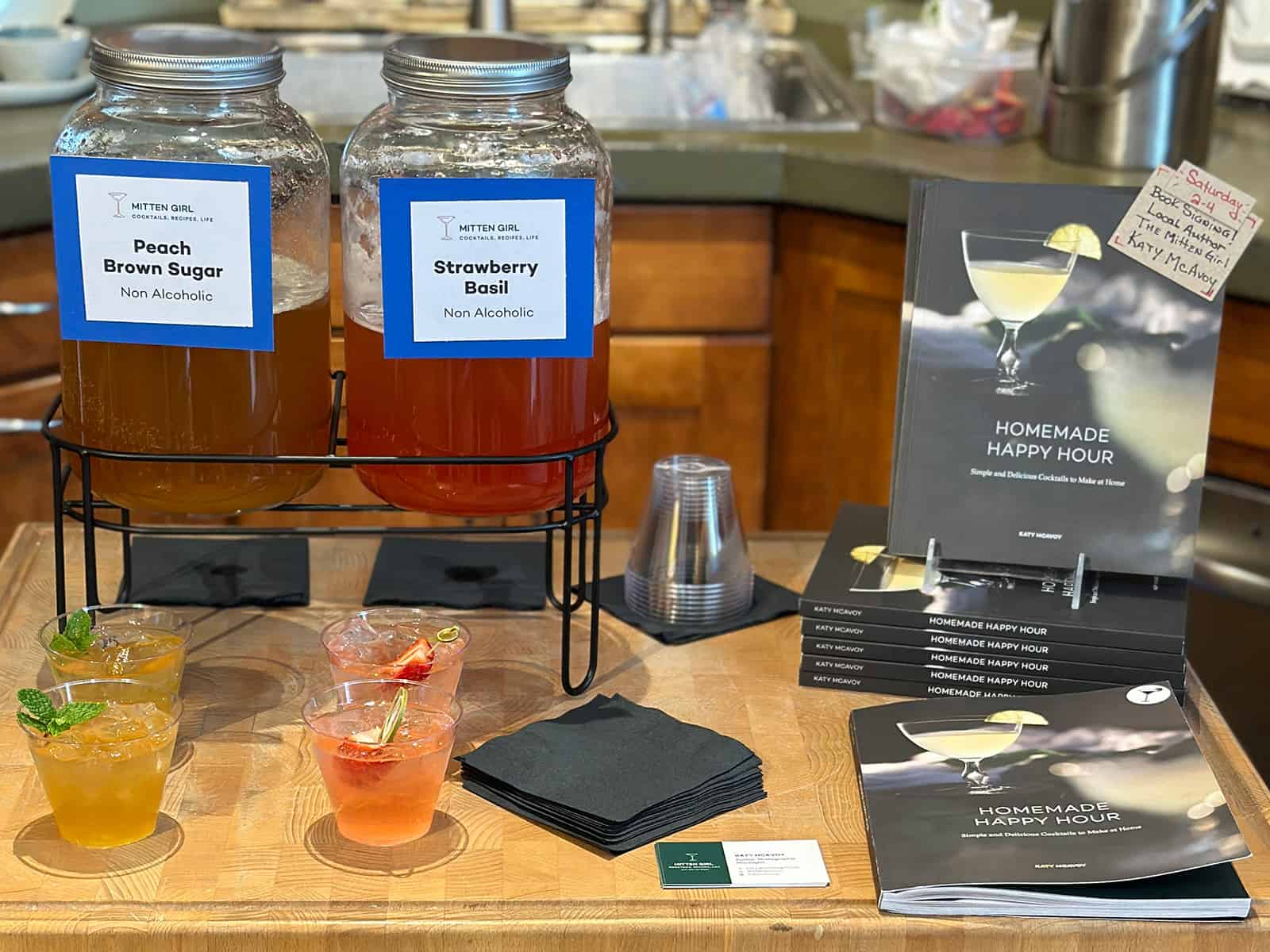 A book signing set up with a display of Homemade Happy Hour books and two pitchers of mocktail samples. 