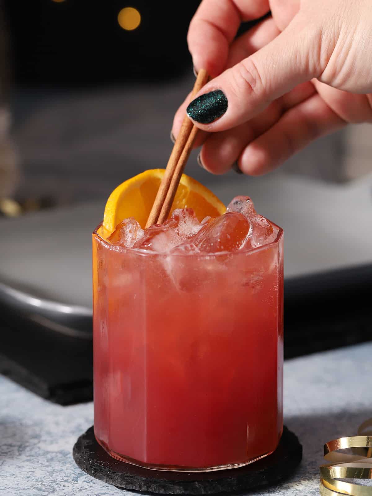 A hand garnishing a glass of cherry holiday mocktail garnished with orange slices and cinnamon sticks.
