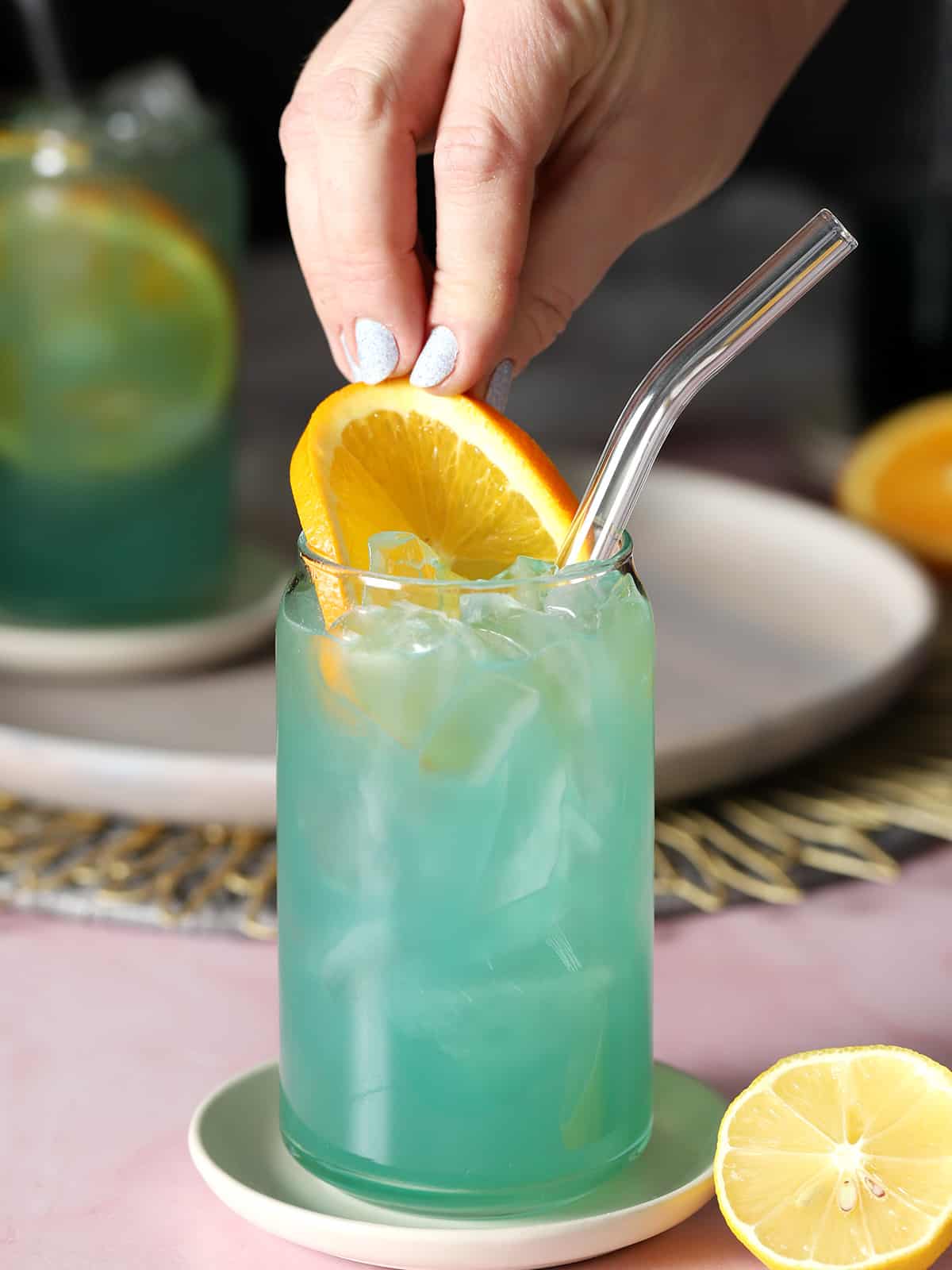 A hand pushing an orange slice into a glass of blue lagoon mocktail.