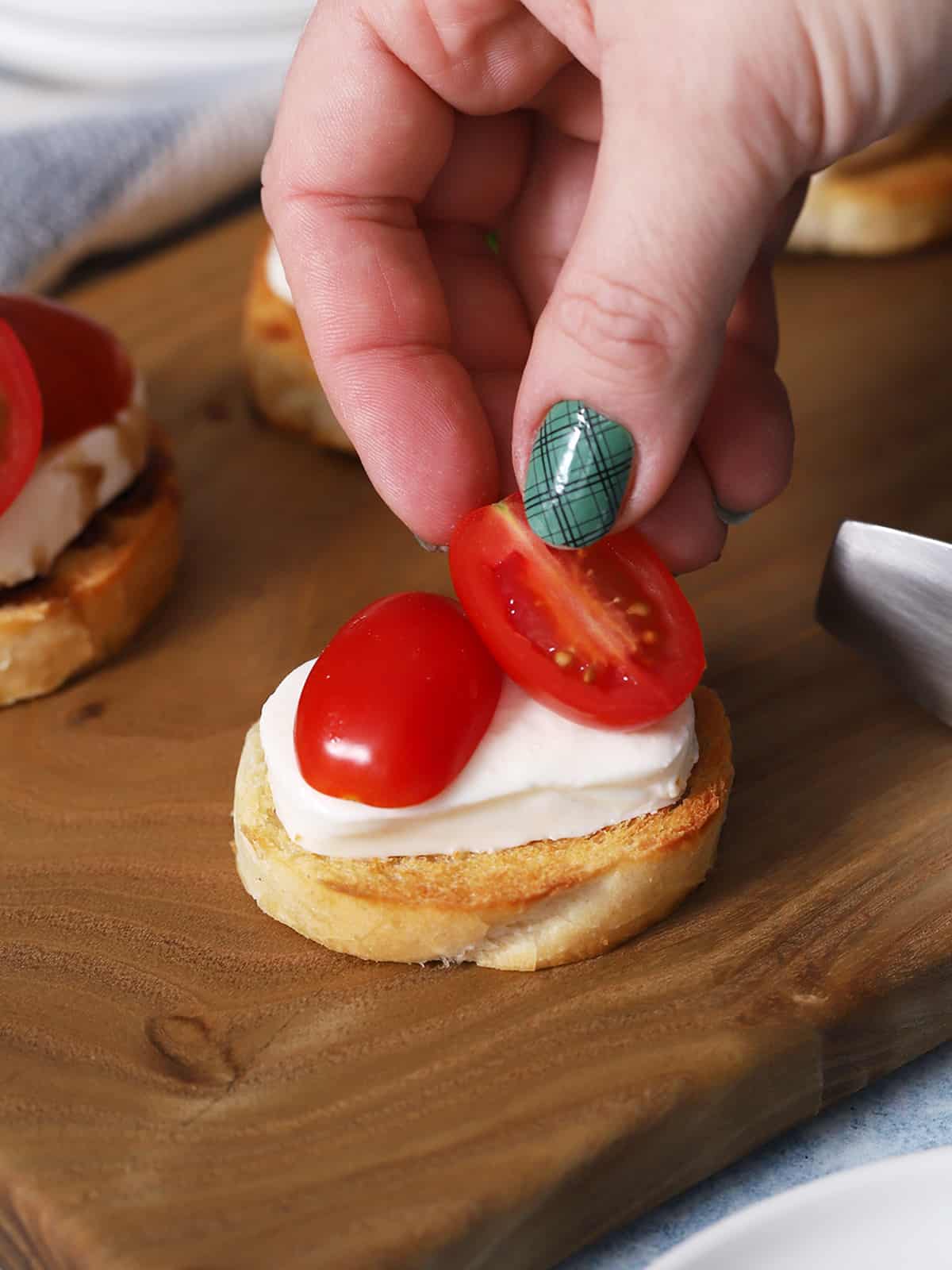 A hand placing half of a cherry tomato onto a crostini. 