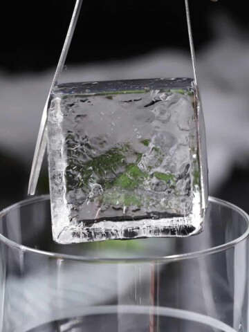 A clear ice cube being lowered into a glass with a pair of tongs.