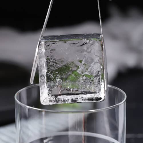 A clear ice cube being lowered into a glass with a pair of tongs.