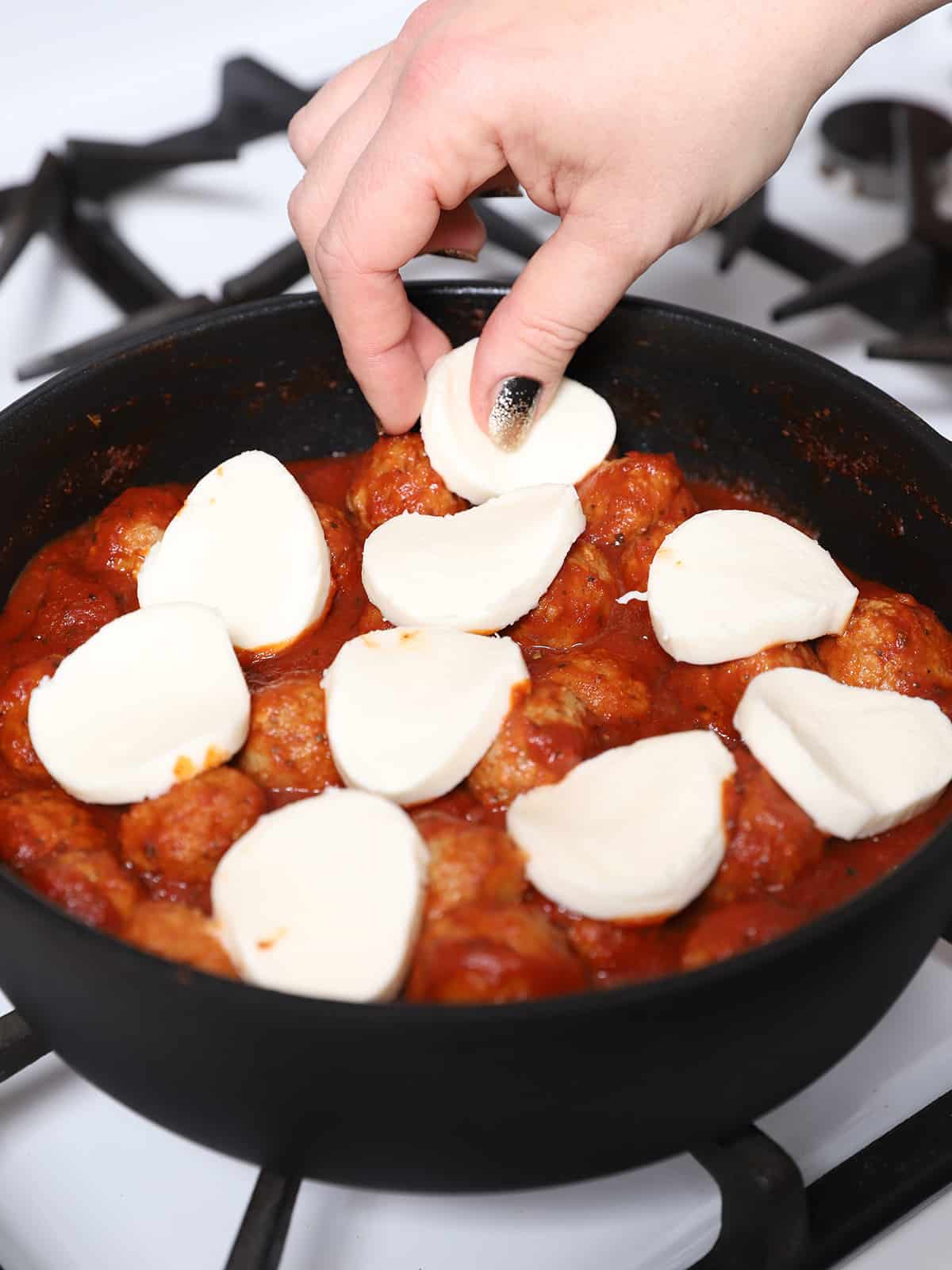A hand laying slices of mozzarella cheese onto the meatballs. 