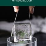A clear ice cube being lowered into a glass with a pair of tongs.