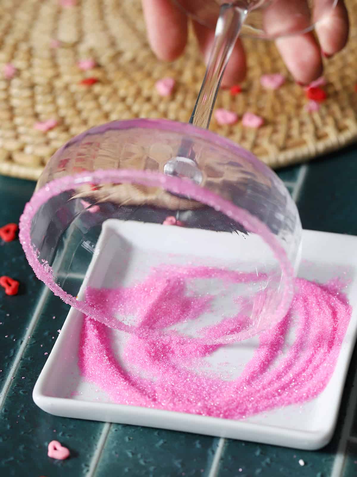 A hand dipping the rim of a glass in bright pink decorating sugar. 