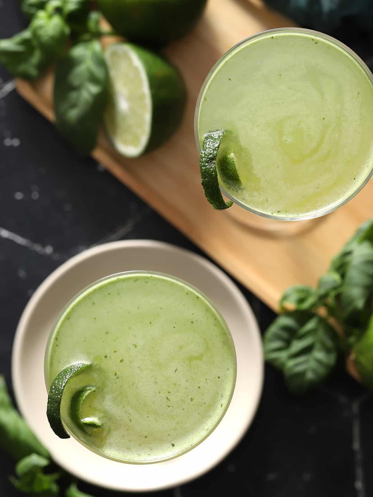 Looking down at two glasses of basil gimlet cocktail garnished with curls of lime peel, surrounded by fresh limes and basil, on cream colored coasters.