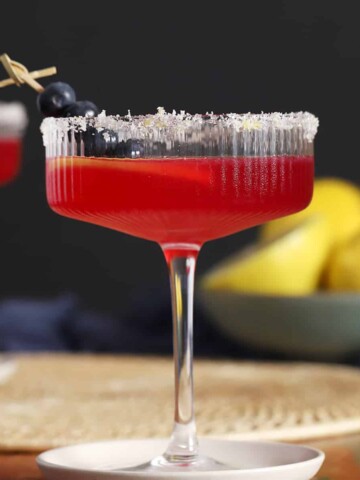 A coupe glass of blueberry lemon drop martini with a lemon sugar rim and cocktail pick of blueberries in the glass.