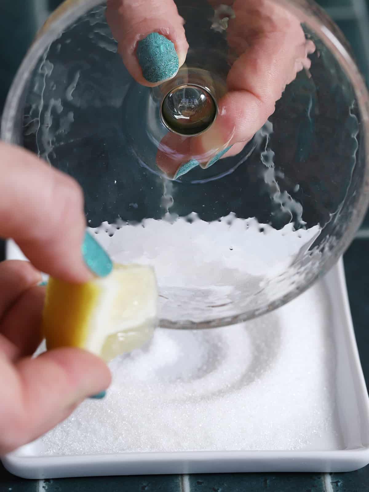 A hand running a slice of lemon around the rim of a coupe glass. 