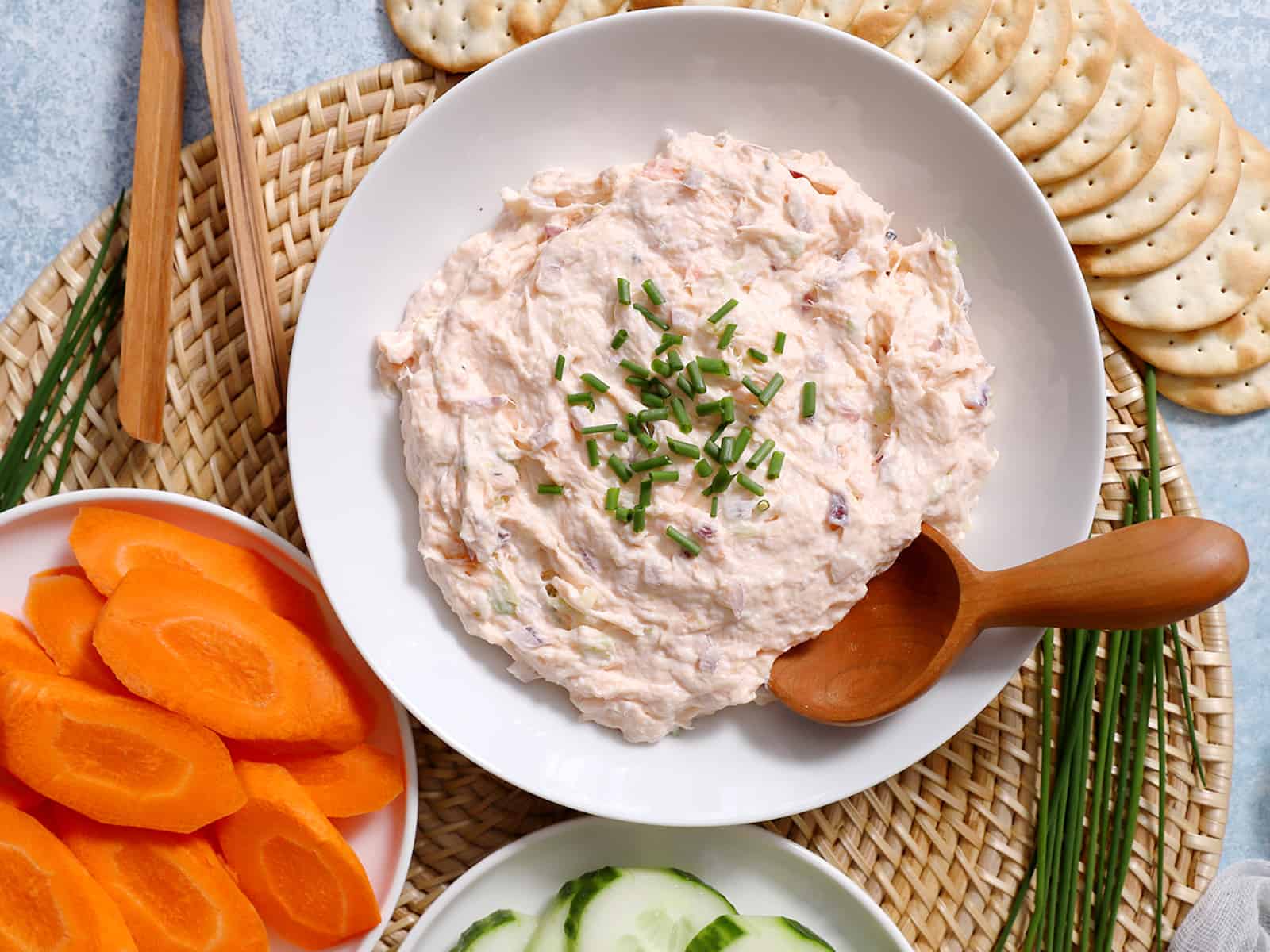 A bowl of smoked salmon dip with a serving spoon in it, surrounded by veggies and crackers.