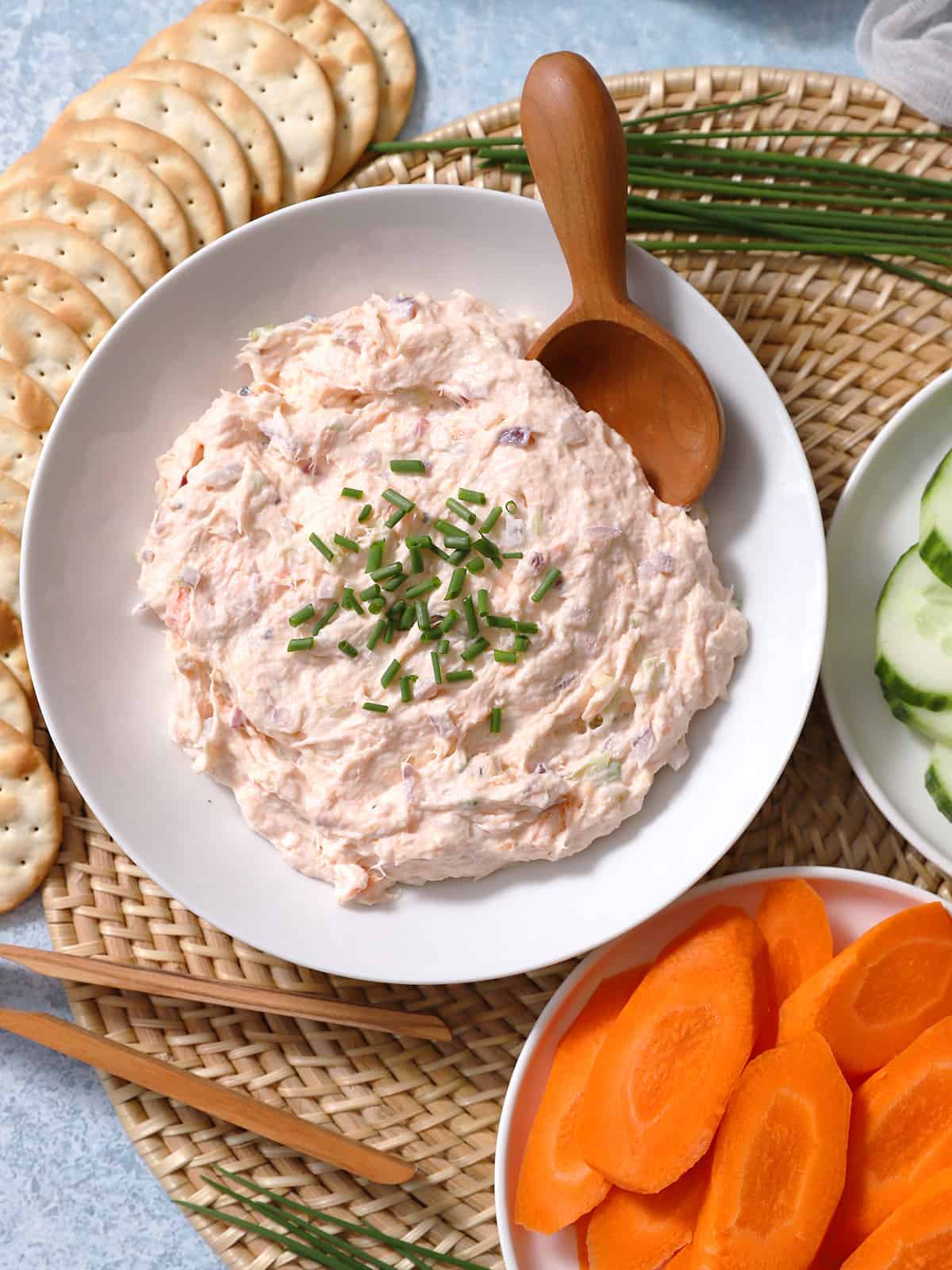 A bowl of smoked salmon dip with a wooden spoon in it surrounded by crackers and raw veggies.