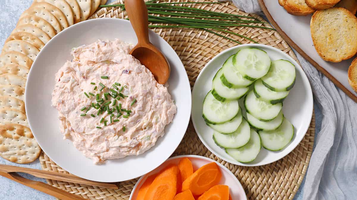 A bowl of smoked salmon dip surrounded by crackers, carrot slices, and cucumber slices. 