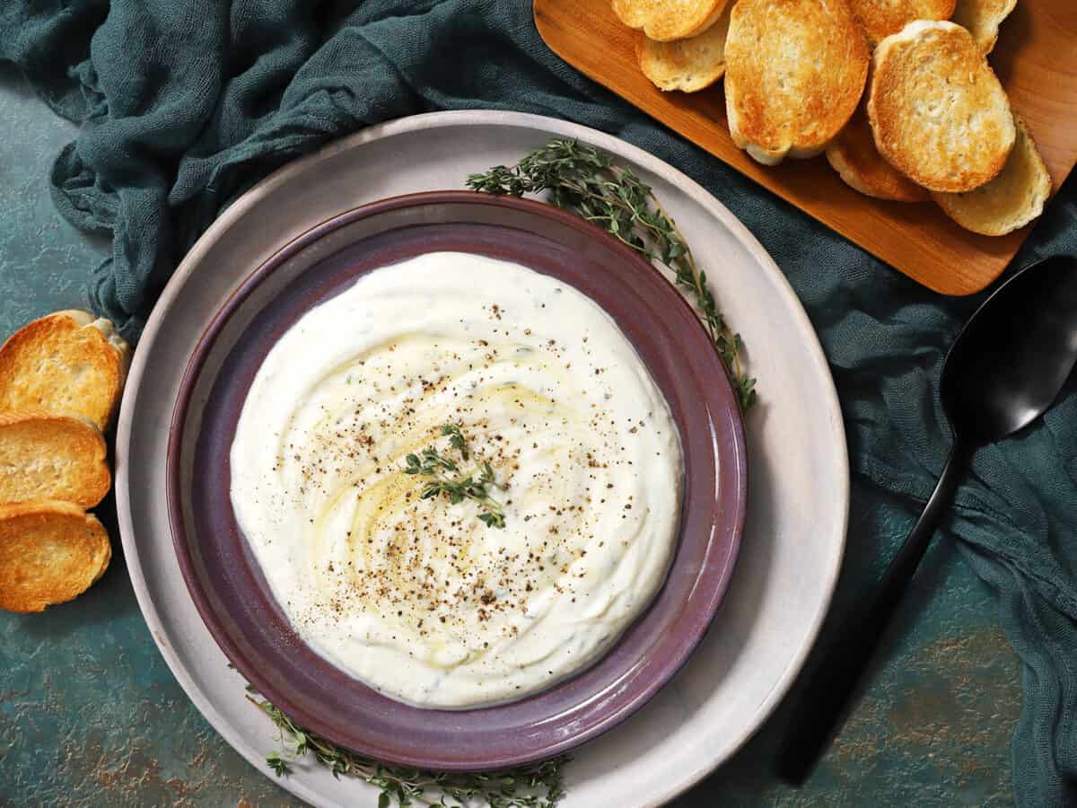 A plate of whipped ricotta dip topped with cracked black pepper, fresh thyme leaves, and swirls of honey.