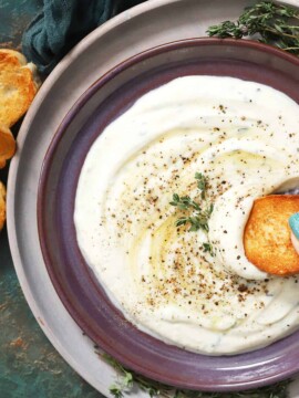 A hand dipping a piece of crostini in a plate of whipped ricotta dip.