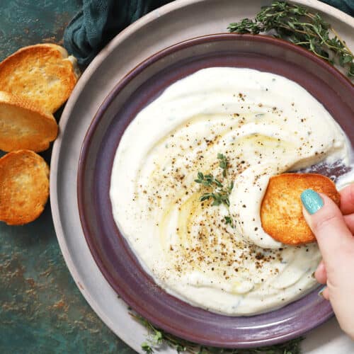 A hand dipping a piece of crostini in a plate of whipped ricotta dip.