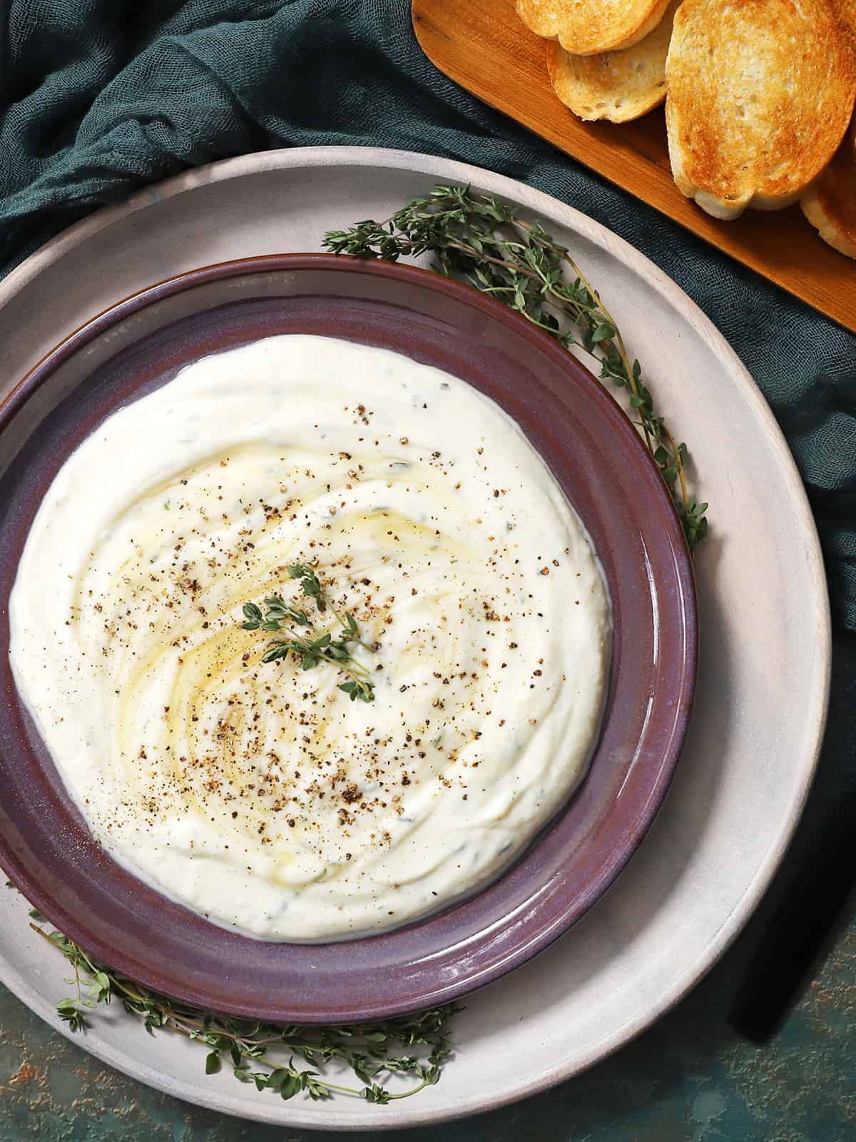 A plate of whipped ricotta dip topped with cracked black pepper, fresh thyme leaves, and swirls of honey. 