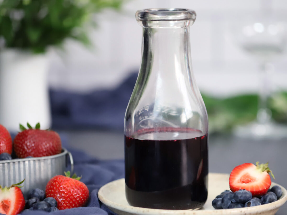 A jar of mixed berry syrup sitting on a small plate with strawberries and blueberries.