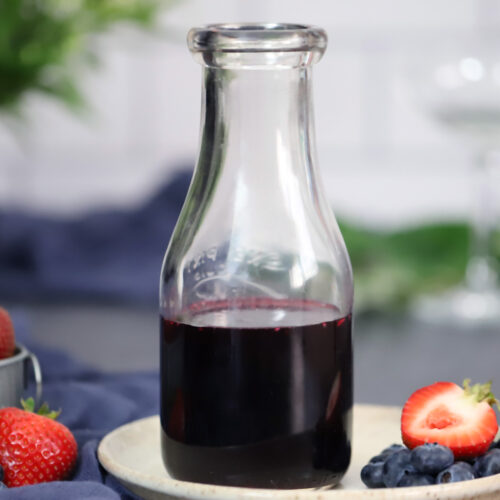 A jar of mixed berry syrup sitting on a small plate with strawberries and blueberries.