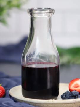 A jar of mixed berry syrup sitting on a small plate with strawberries and blueberries.