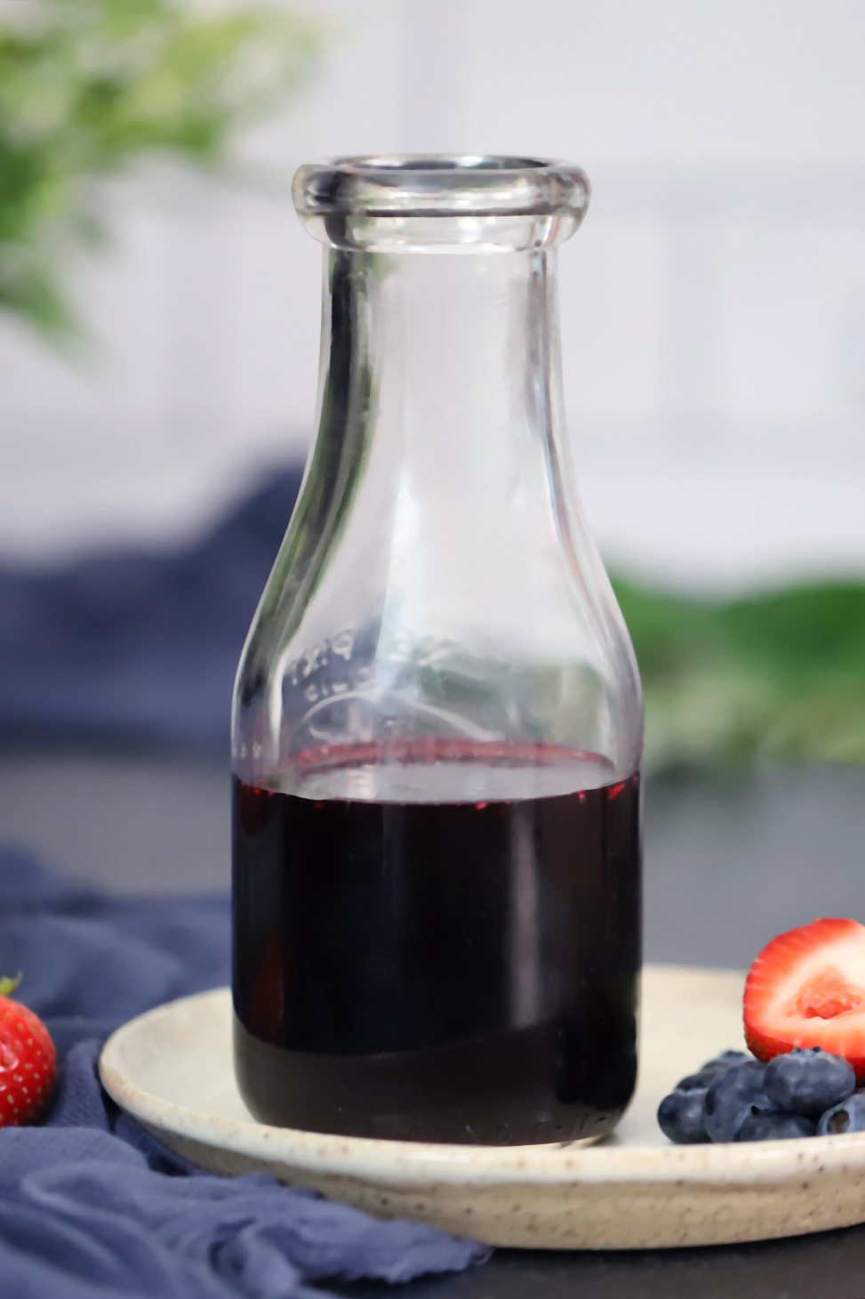 A jar of mixed berry syrup sitting on a small plate with strawberries and blueberries.