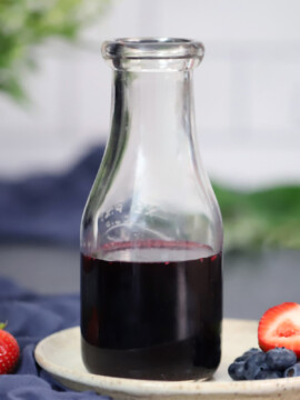 A jar of mixed berry syrup sitting on a small plate with strawberries and blueberries.