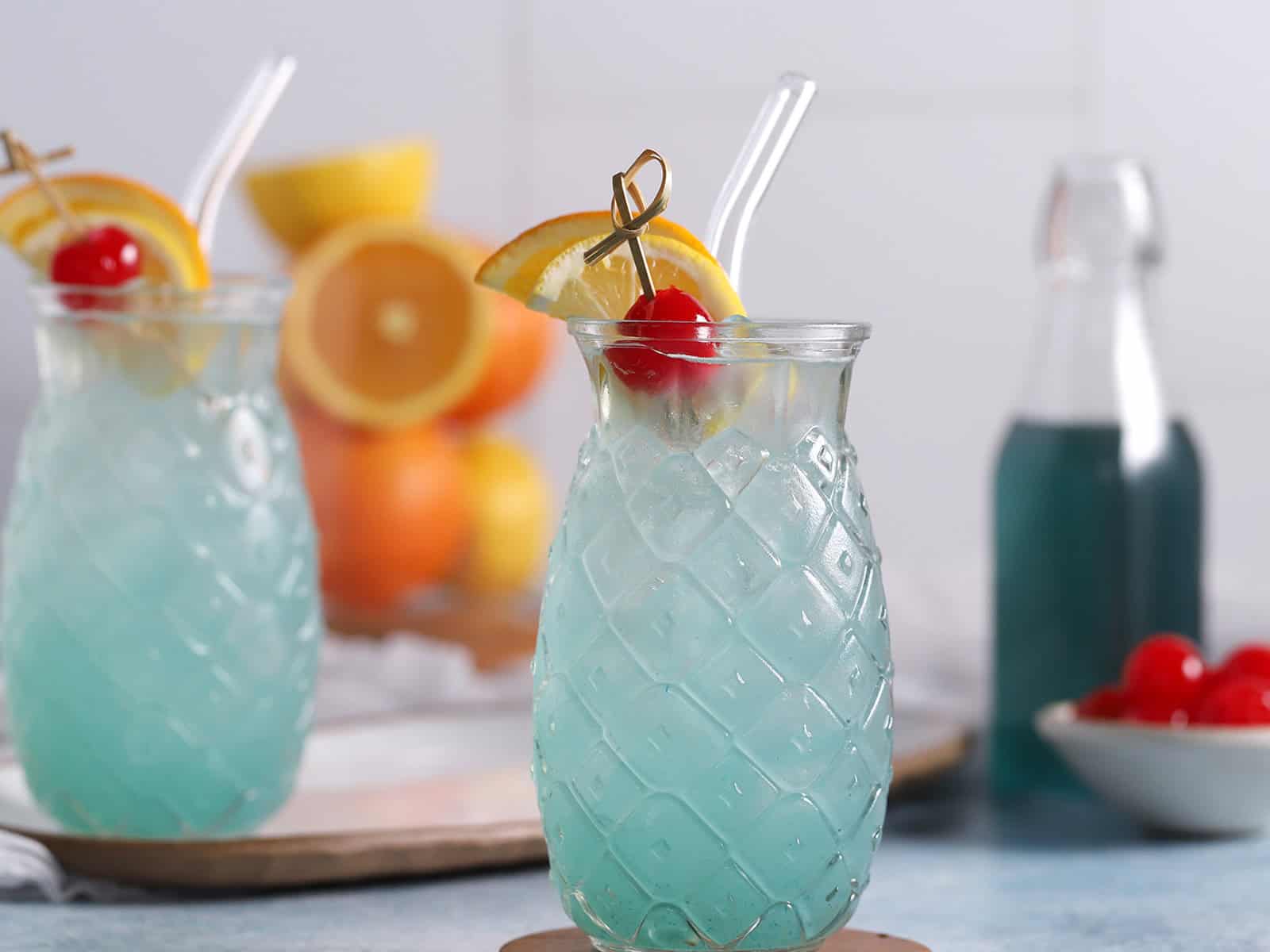 Glasses of blue lagoon cocktail garnished with oranges and cherries, with a jar of oranges in the background.