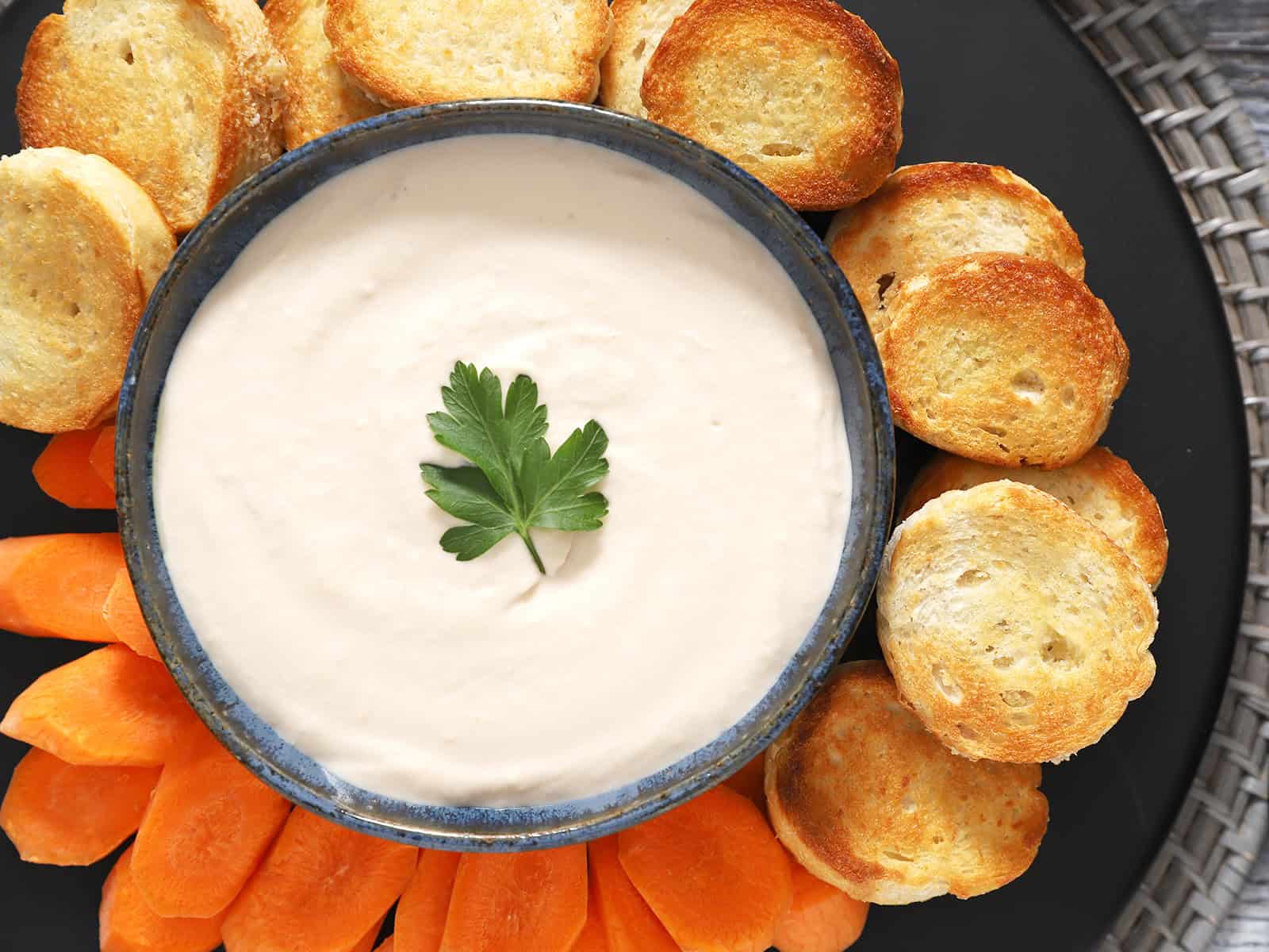 A bowl of white bean spread surrounded by crostini and carrots.