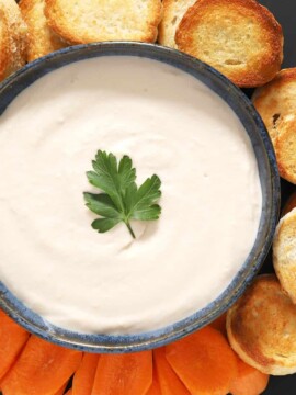 A bowl of white bean spread surrounded by crostini and carrots.