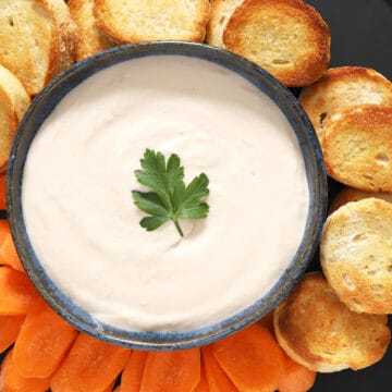 A bowl of white bean spread surrounded by crostini and carrots.