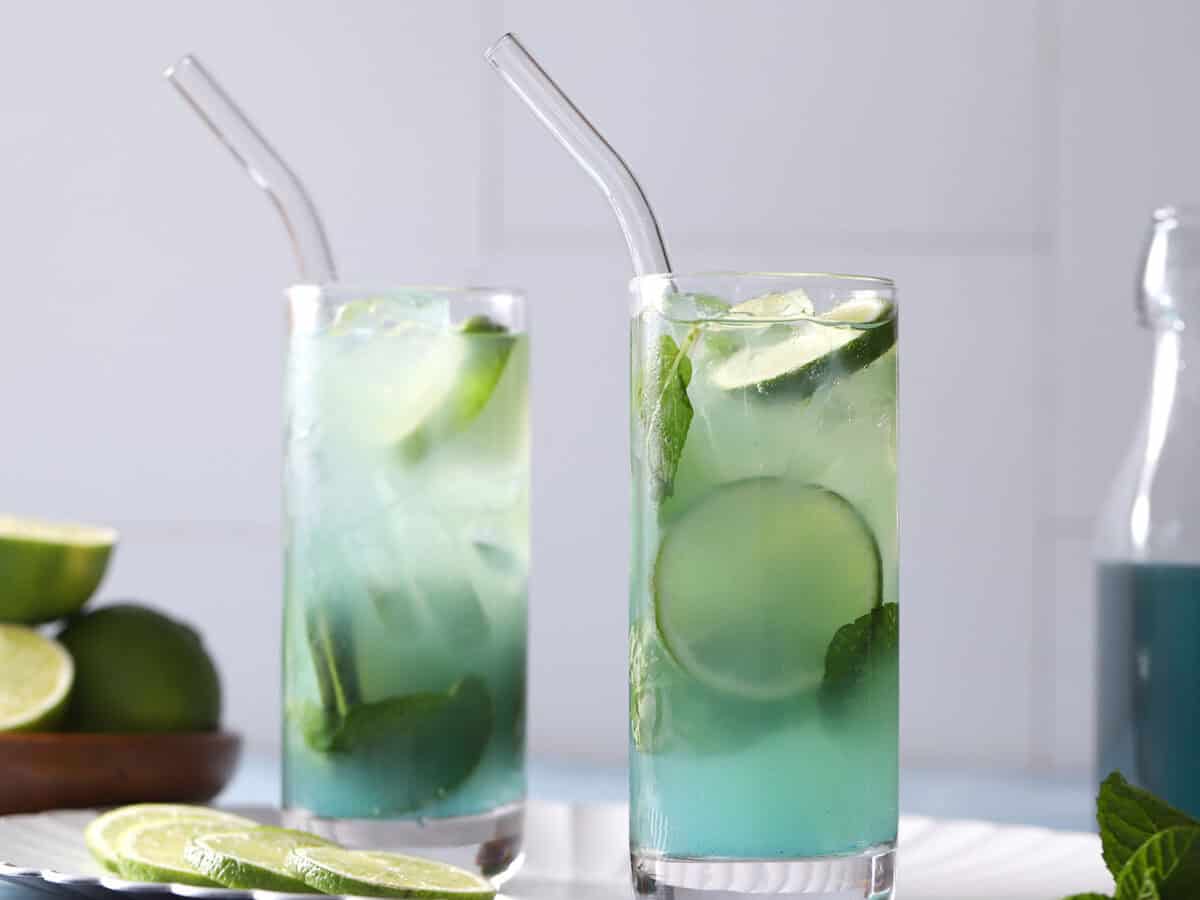 Two glasses of blue mojito cocktails next to a jar of blue curaƧao syrup.