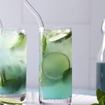 Two glasses of blue mojito cocktails next to a jar of blue curaçao syrup.