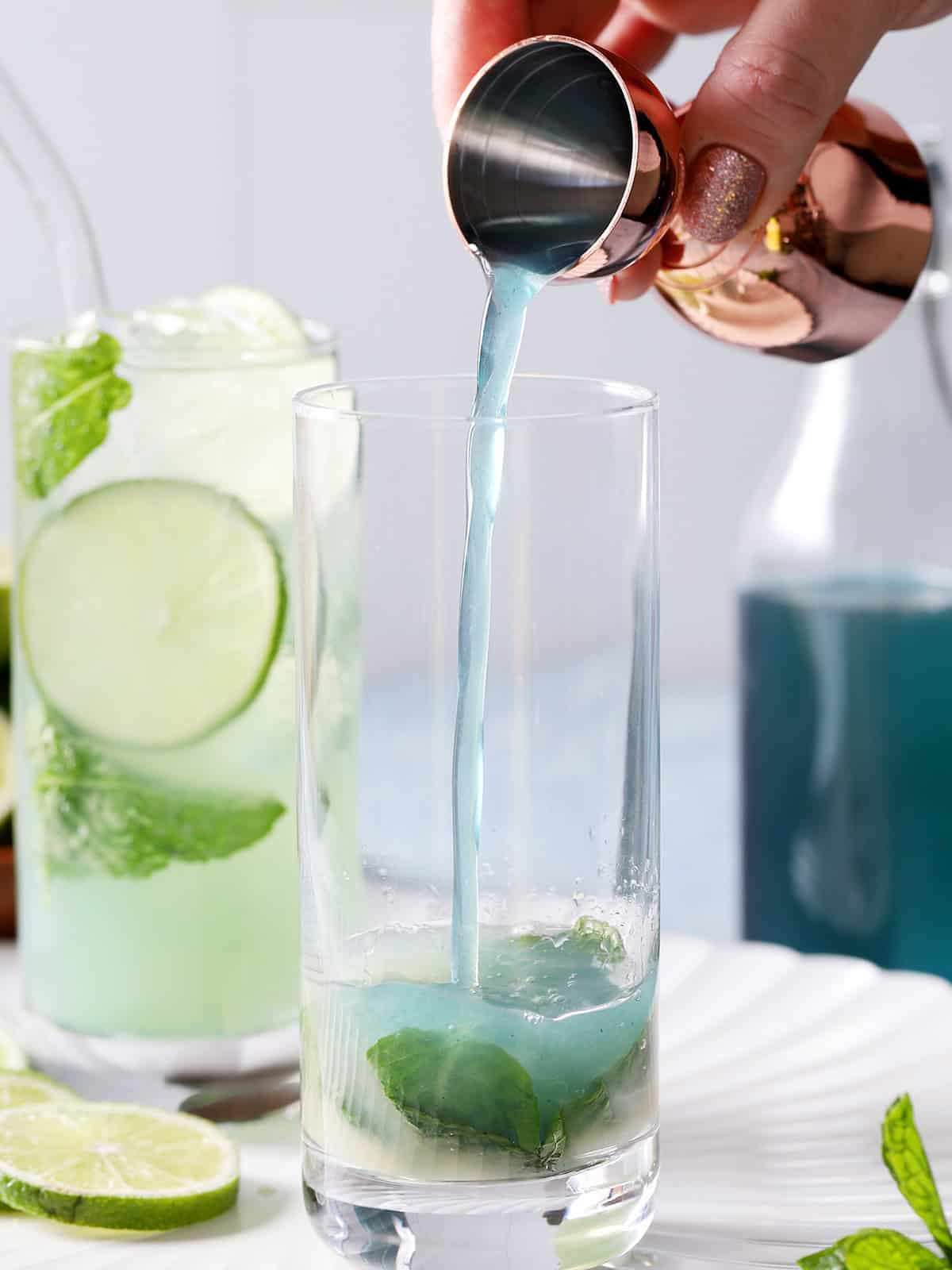 Blue curaçao syrup being poured into a tall glass with mint and lime juice in it. 