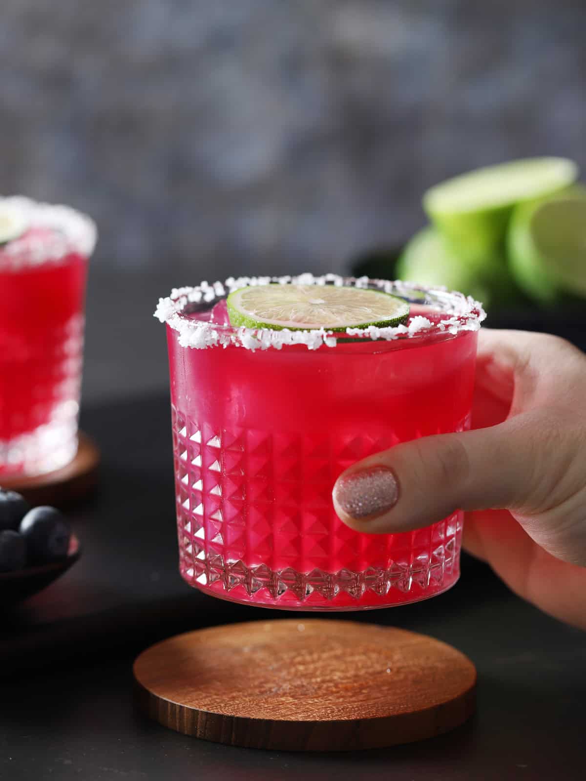 A hand lifting up a glass filled with blueberry margarita. 