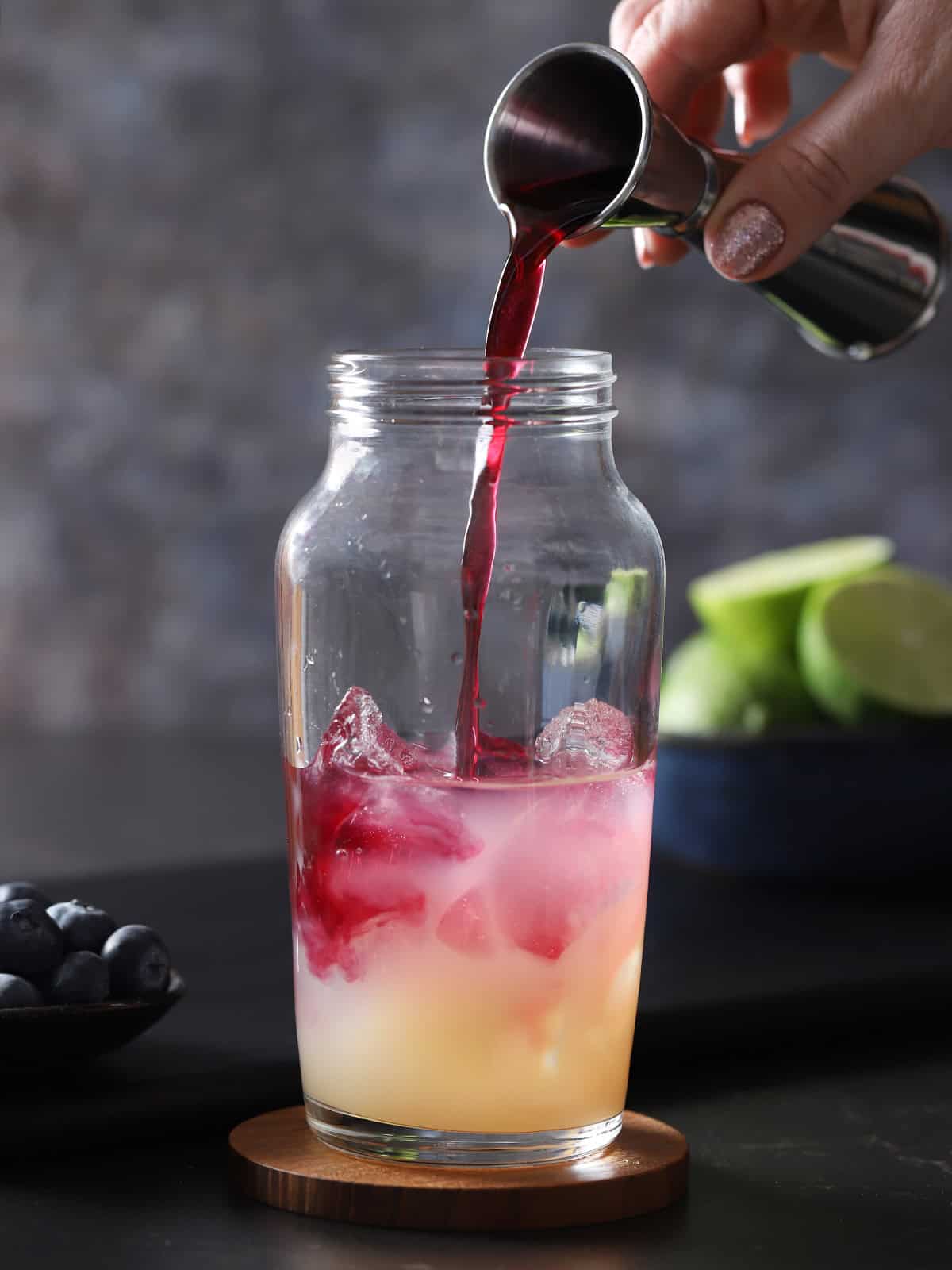 Blueberry syrup being poured from a jigger into a cocktail shaker filled with lime juice, tequila, and ice. 