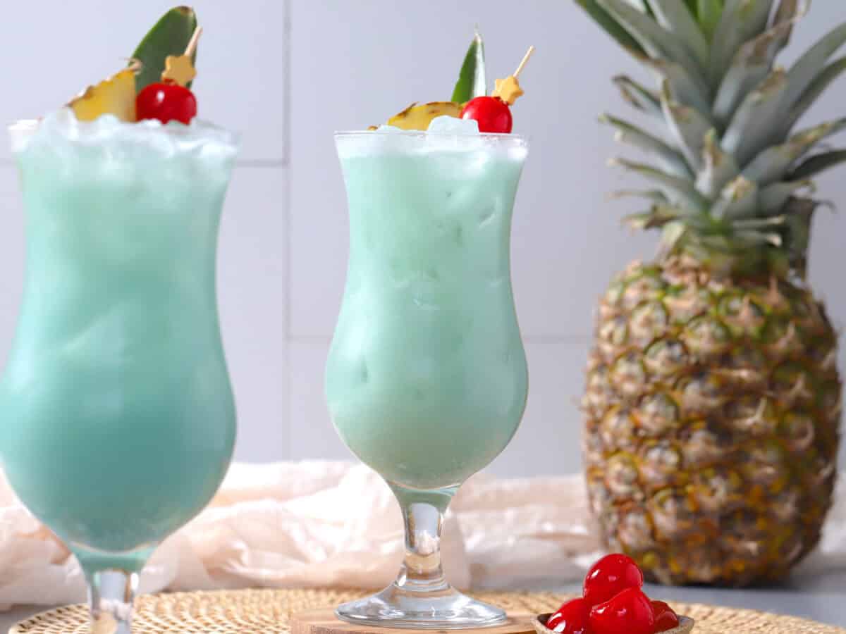 A pair of hurricane glasses filled with blue hawaiian cocktail with a pineapple in the background.