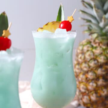 A pair of hurricane glasses filled with blue hawaiian cocktail with a pineapple in the background.