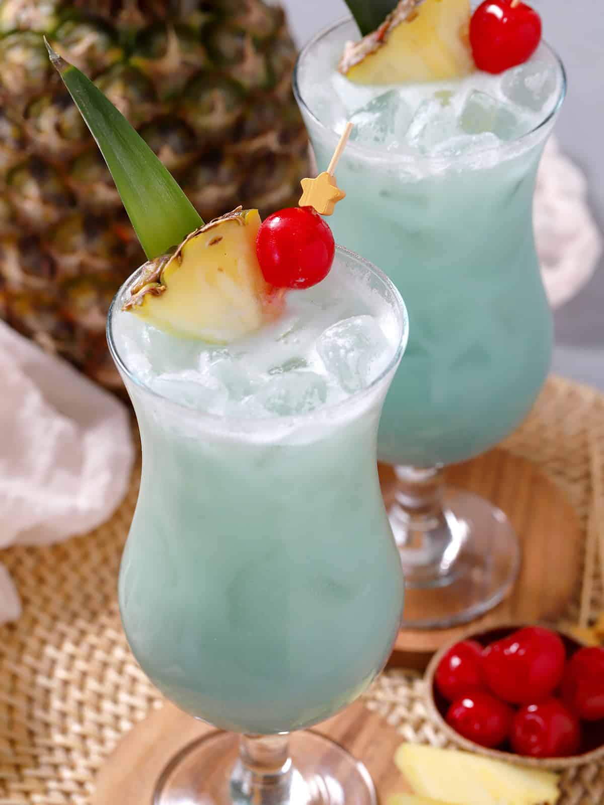 A pair of hurricane glasses filled with blue hawaiian cocktail with a pineapple in the background.