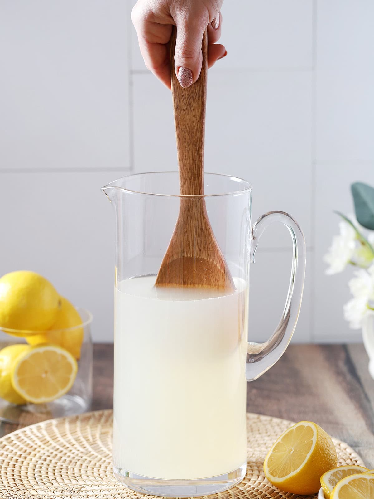 A hand using a large wooden spoon to stir a pitcher of lemonade.