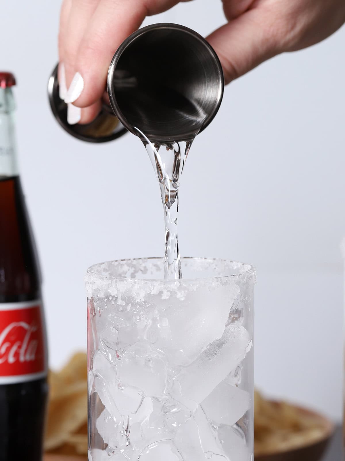 Tequila being poured from a jigger into a tall glass with ice.