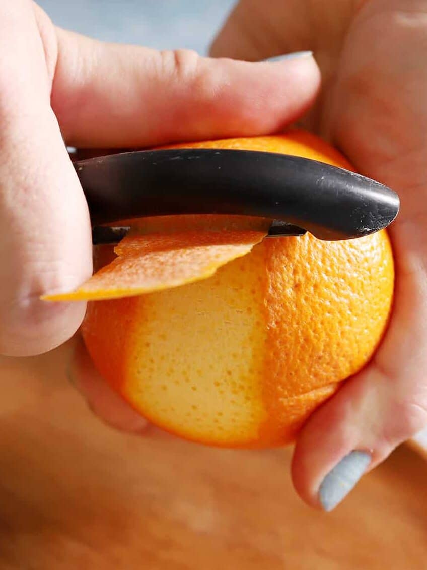 A hand using a vegetable peeler to peel an orange.