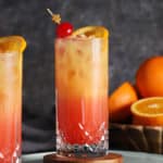 A glass of tequila sunrise cocktail garnished with cherries and fresh orange.