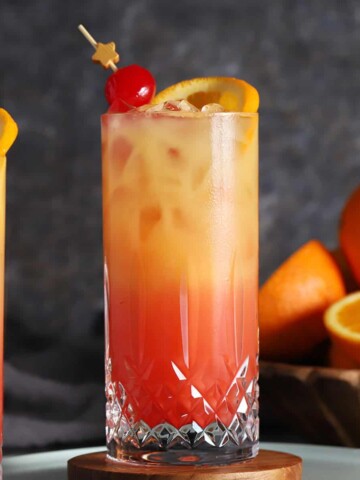 A glass of tequila sunrise cocktail garnished with cherries and fresh orange.