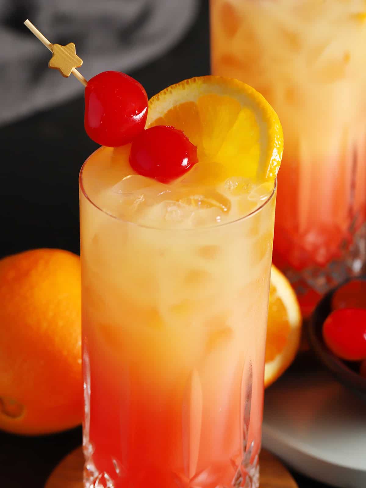 A glass of tequila sunrise cocktail garnished with cherries and fresh orange.