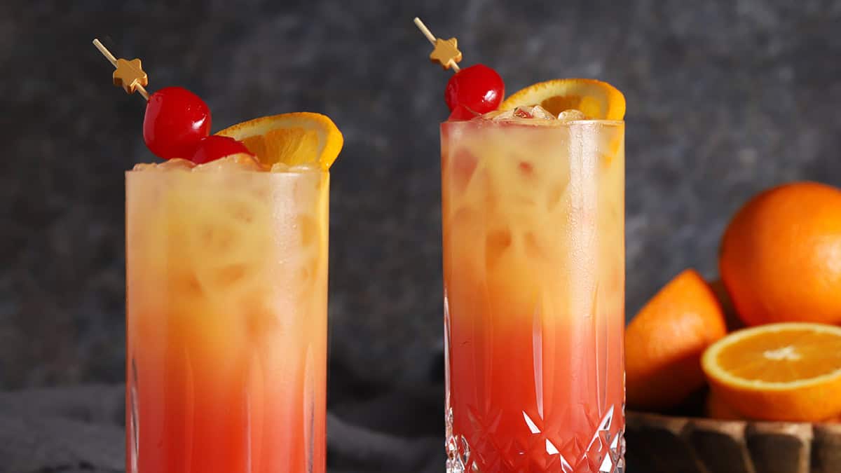 Glasses of tequila sunrise cocktail garnished with cherries and fresh orange.