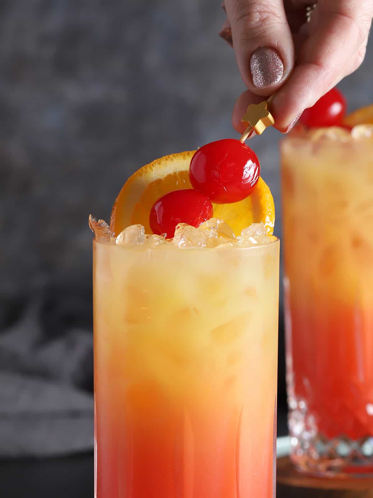 A hand placing a cocktail pick of maraschino cherries into a glass of tequila sunrise cocktail.