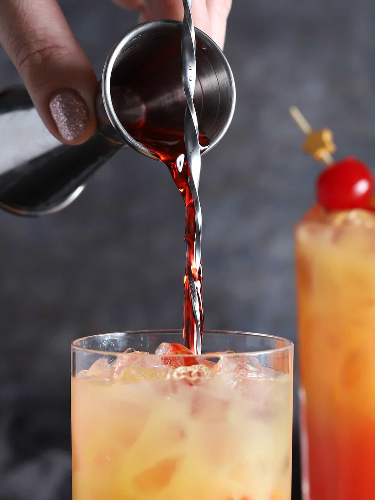 A hand pouring grenadine syrup from a jigger down the handle of a bar spoon set into a glass.